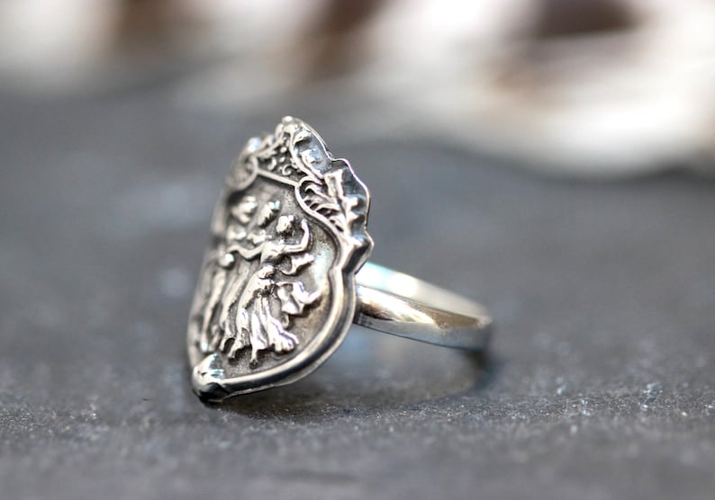 Puede incluir: Un anillo de plata con un dise&ntilde;o de escudo de armas detallado que presenta tres figuras. El anillo est&aacute; hecho de plata de ley y tiene un acabado pulido.
