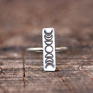 May include: A silver ring with a vertical rectangular band featuring a design of eight moon phases.