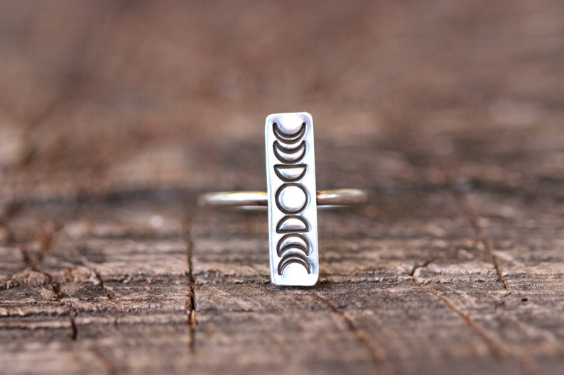 May include: A silver ring with a vertical rectangular band featuring a design of eight moon phases.