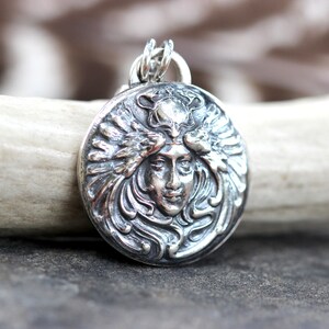 May include: Silver pendant featuring a detailed, ornate design of a woman's face with flowing hair and wings. The pendant is circular and has a textured surface.