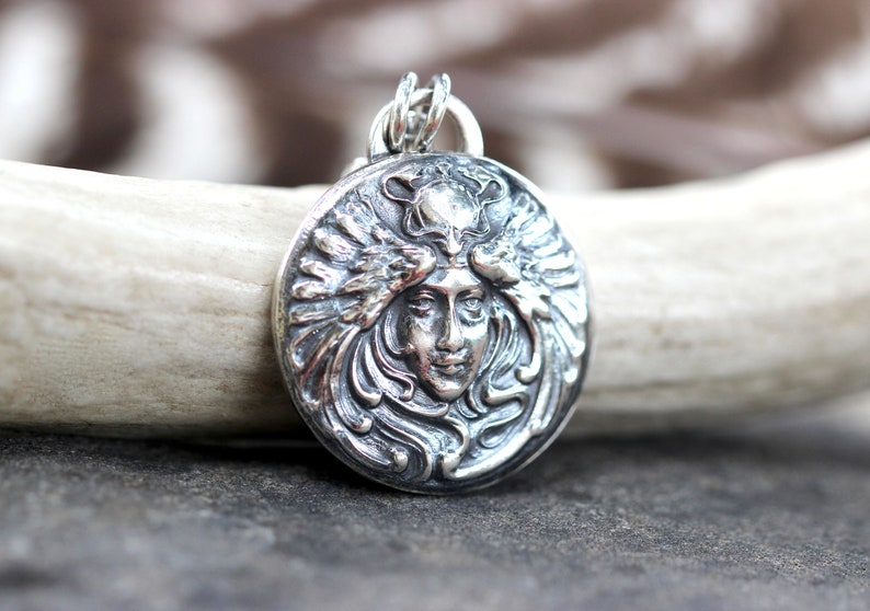 May include: Silver pendant featuring a detailed, ornate design of a woman's face with flowing hair and wings. The pendant is circular and has a textured surface.