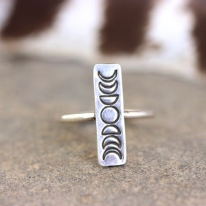May include: A silver ring with a rectangular band featuring a design of eight crescent moons in different phases.