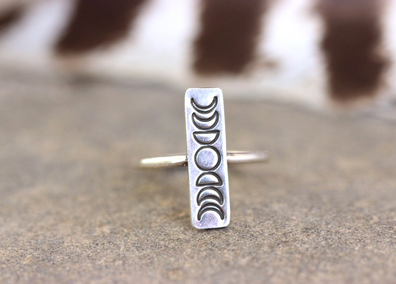 May include: A silver ring with a rectangular band featuring a design of eight crescent moons in different phases.