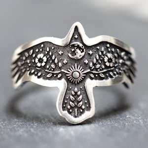 May include: A silver-toned ring featuring an embossed bird with wings spread wide. The design includes intricate engravings of a crescent moon, a sun, small stars, floral patterns, and leafy branches across the surface of the metal band.