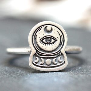Puede incluir: Anillo de plata con un diseño detallado de una bola de cristal con un ojo, una luna creciente y tres lunas crecientes. El anillo tiene un acabado pulido y una banda fina. Una joya única para los interesados en la astrología y el ocultismo.