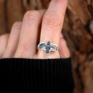 May include: A silver ring featuring a detailed bird design. The ring's focal point is a stylized bird with outstretched wings, accented with intricate feather detailing. The ring is worn on a finger, showcasing the craftsmanship and design.