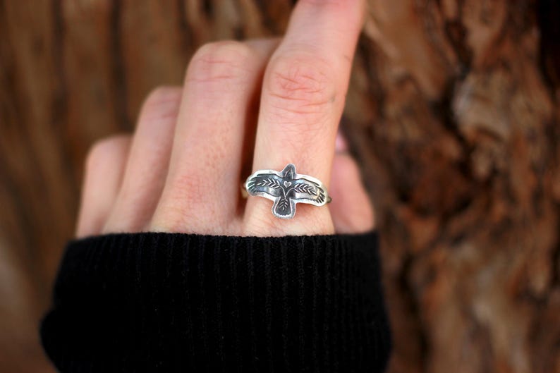 May include: A silver ring featuring a detailed bird design. The ring's focal point is a stylized bird with outstretched wings, accented with intricate feather detailing. The ring is worn on a finger, showcasing the craftsmanship and design.