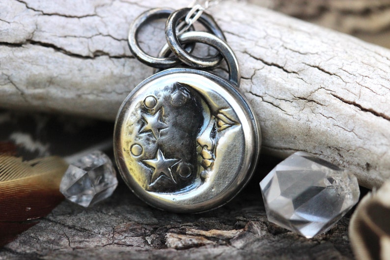 Puede incluir: Un colgante de plata con una luna creciente con tres estrellas y una superficie texturizada. El colgante est&aacute; unido a una cadena de plata.