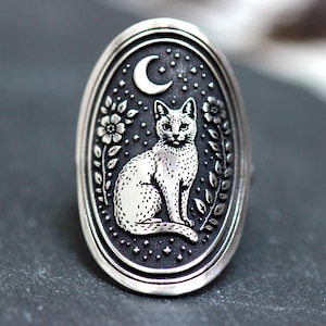 May include: Silver-toned ring with an oval face featuring a detailed cat design. The cat sits beneath a crescent moon and stars, surrounded by floral and leaf motifs. The ring has a detailed, vintage aesthetic.