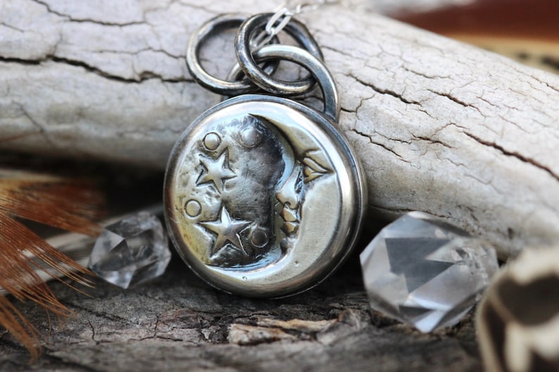 Puede incluir: Un colgante de plata con una luna creciente con una cara y tres estrellas. El colgante est&aacute; en una cadena de plata y est&aacute; descansando sobre un trozo de madera flotante.