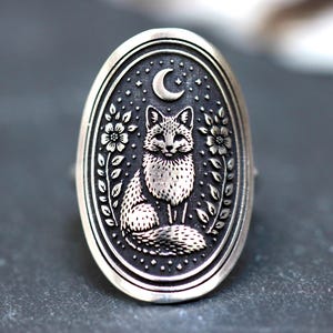 Könnte beinhalten: Silberfarbener Ring mit ovaler Oberfläche, verziert mit einem detaillierten Fuchsmotiv. Der Fuchs sitzt unter einem Mond und Sternen, umgeben von floralen und Blatt-Akzenten. Der Ring hat eine polierte Oberfläche.