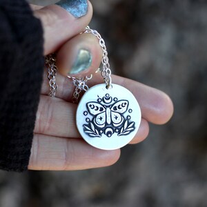 May include: A silver necklace with a round pendant featuring a black and white illustration of a moth with moon and stars. The pendant is hanging from a silver chain.
