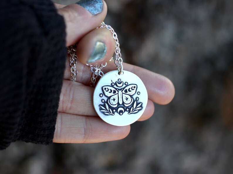 May include: A silver necklace with a round pendant featuring a black and white illustration of a moth with moon and stars. The pendant is hanging from a silver chain.
