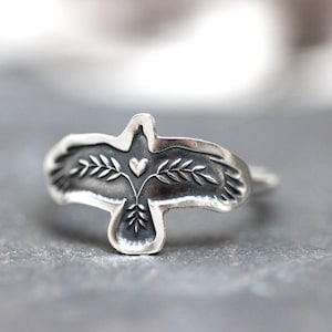 May include: A silver ring featuring a stylized bird design. The bird has outstretched wings, with a heart and leaf details. The ring is made of a shiny, metallic material, and the design is slightly raised.