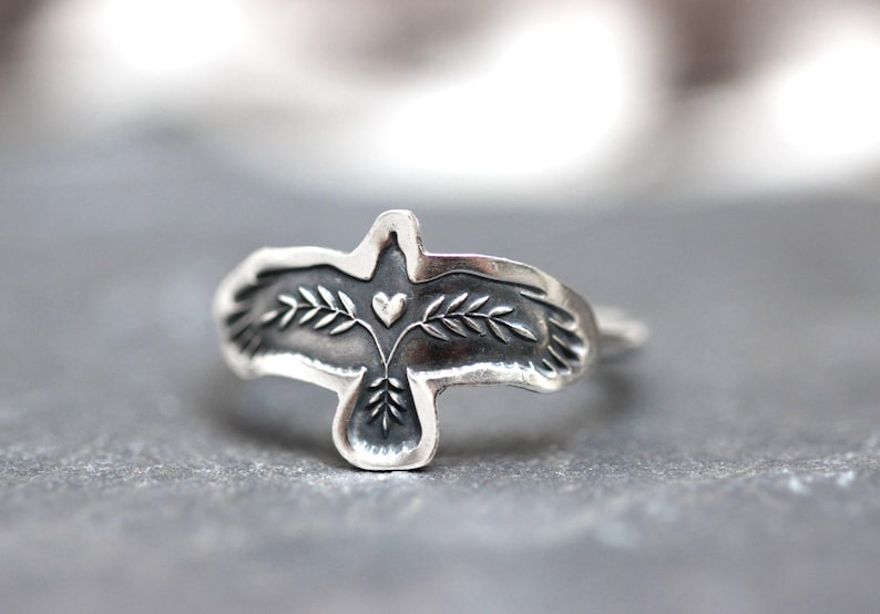 May include: A silver ring featuring a stylized bird design. The bird has outstretched wings, with a heart and leaf details. The ring is made of a shiny, metallic material, and the design is slightly raised.