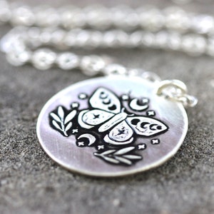 May include: A silver necklace with a round pendant featuring a black and white etched design of a moth with crescent moons and stars.