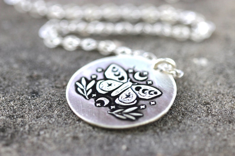 May include: A silver necklace with a round pendant featuring a black and white etched design of a moth with crescent moons and stars.