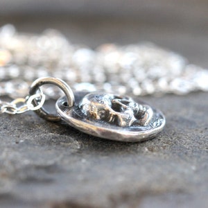 May include: A silver pendant necklace featuring a detailed skull design. The pendant is round and has a textured surface. The necklace chain is also silver and is visible in the background. The jewelry is displayed on a gray surface.