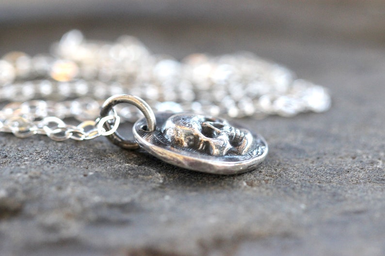 May include: A silver pendant necklace featuring a detailed skull design. The pendant is round and has a textured surface. The necklace chain is also silver and is visible in the background. The jewelry is displayed on a gray surface.