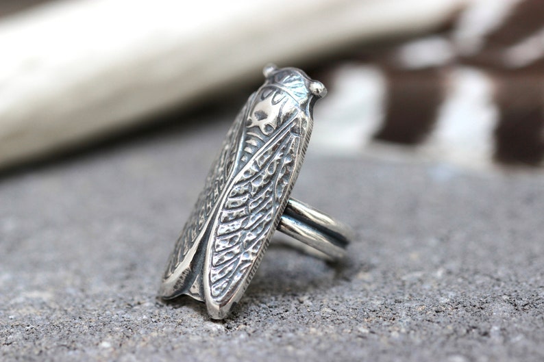 May include: A silver ring featuring a detailed cicada design. The ring is made of sterling silver and has a textured finish.