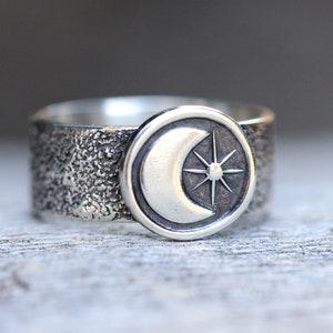 May include: A silver ring with a textured band and a round, raised design featuring a crescent moon and a star.