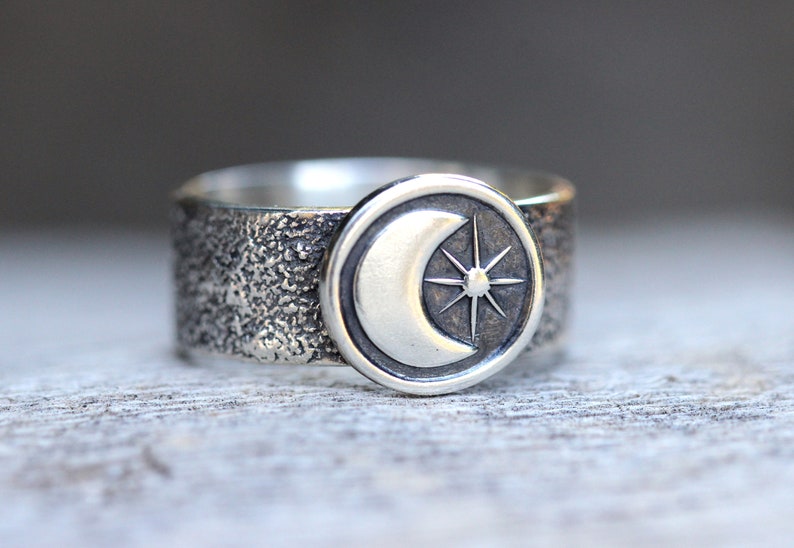 May include: A silver ring with a textured band and a round, raised design featuring a crescent moon and a star.