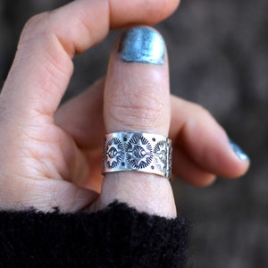 May include: A wide, silver-toned ring with an intricate pattern of sun-like designs. The ring is worn on a finger, showcasing its detailed craftsmanship. The ring's design features repeating circular patterns with radiating lines.