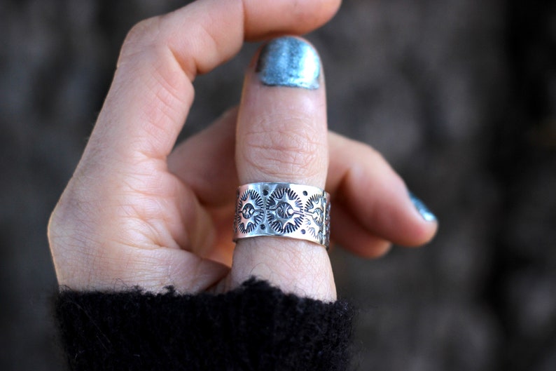 May include: A wide, silver-toned ring with an intricate pattern of sun-like designs. The ring is worn on a finger, showcasing its detailed craftsmanship. The ring's design features repeating circular patterns with radiating lines.