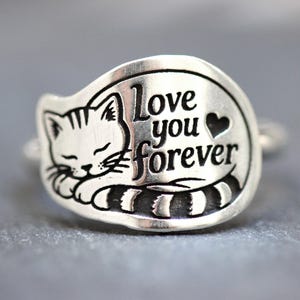 May include: A silver-toned ring featuring an engraved design of a sleeping cat with a striped tail. The text "Love you forever" is etched in black lettering next to the cat, accompanied by a small heart symbol on the right side of the band.