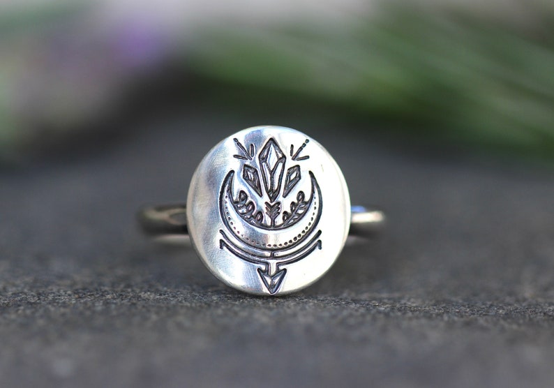 May include: A silver ring with a round face featuring an etched design of a crescent moon, a crystal, and a stylized arrow.