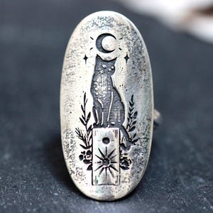 May include: An oval-shaped silver ring featuring an engraved design of a cat sitting on a rectangular pedestal. Above the cat is a crescent moon and stars, while below the pedestal is a sun symbol surrounded by etched floral and leaf patterns.