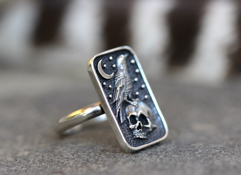 May include: A silver ring with a rectangular design featuring a raven perched on a skull with a crescent moon and stars in the background.