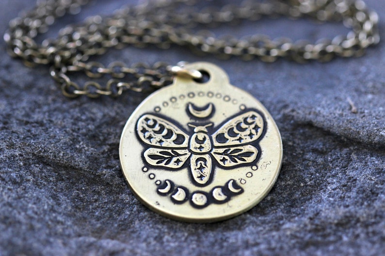 Peut inclure: Un collier en laiton avec un pendentif rond orn&eacute; d'un motif grav&eacute; d&eacute;taill&eacute; d'un papillon de nuit avec des croissants de lune et des &eacute;toiles. Le pendentif est attach&eacute; &agrave; une cha&icirc;ne en laiton.