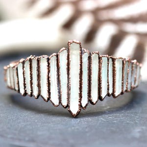 May include: A copper-colored metal bracelet with a series of light green, rectangular crystal-like stones. The stones are set in a geometric pattern, creating a crown-like design. The bracelet is displayed on a gray surface.