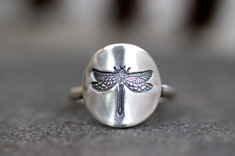 May include: A silver ring with an oval face featuring a detailed dragonfly design. The dragonfly is etched in dark gray against the brushed silver background. The ring is a close-up shot, showcasing the intricate details.