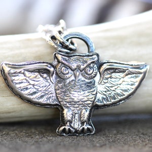 May include: A silver owl pendant with outstretched wings. The owl has detailed feathers and large, round eyes.