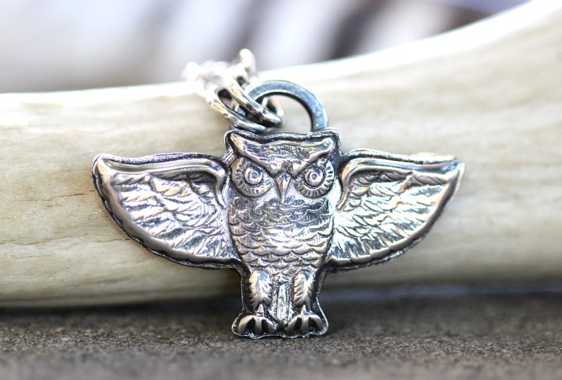 May include: A silver owl pendant with outstretched wings. The owl has detailed feathers and large, round eyes.