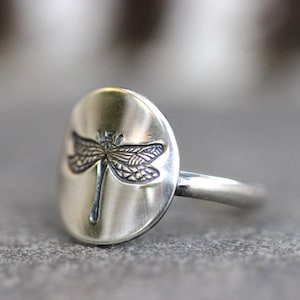 May include: A silver ring featuring a detailed dragonfly design. The dragonfly is etched onto a smooth, oval-shaped face. The ring has a simple band and a brushed metal finish, suitable for everyday wear.