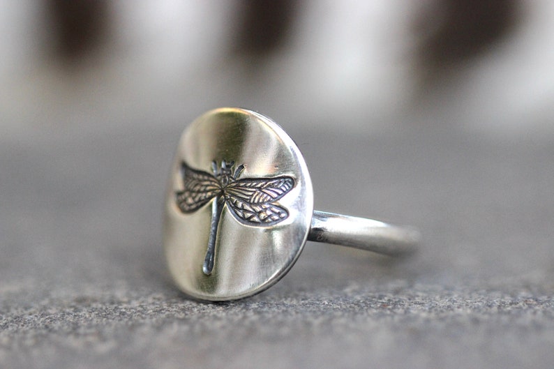 May include: A silver ring featuring a detailed dragonfly design. The dragonfly is etched onto a smooth, oval-shaped face. The ring has a simple band and a brushed metal finish, suitable for everyday wear.