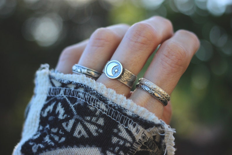 May include: A hand wearing three silver rings. The middle ring features a crescent moon with a star design. The other two rings are plain bands.