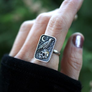 May include: A silver ring with a rectangular design featuring a raven perched on a skull with a crescent moon in the background. The ring is set against a green background.