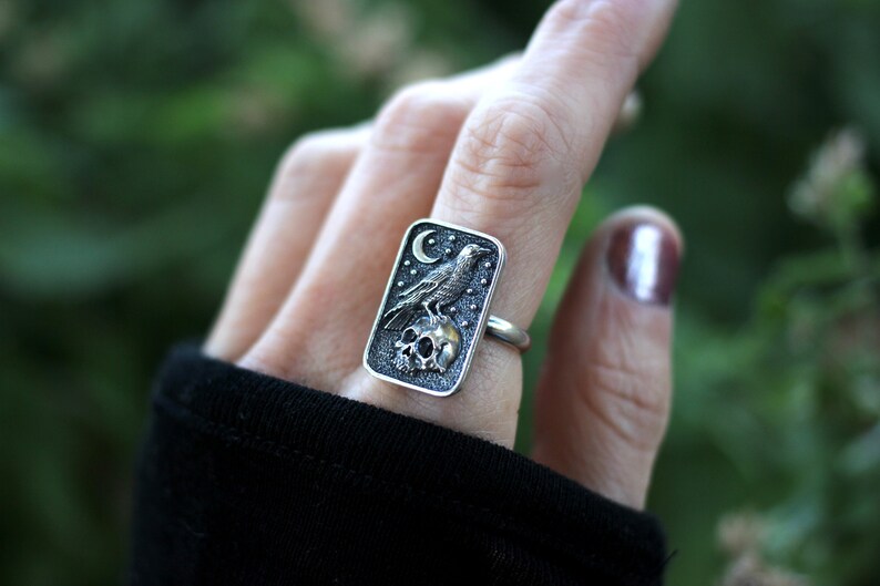 May include: A silver ring with a rectangular design featuring a raven perched on a skull with a crescent moon in the background. The ring is set against a green background.