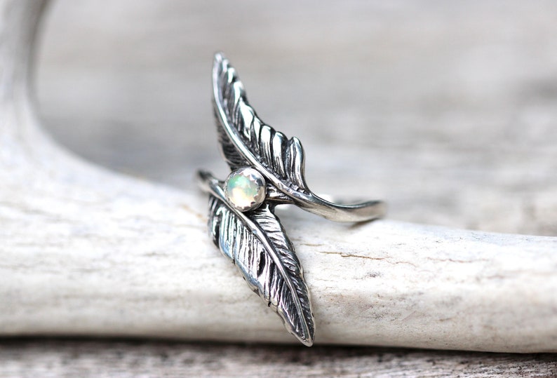 May include: A silver ring with two feather designs and a white opal gemstone in the center.