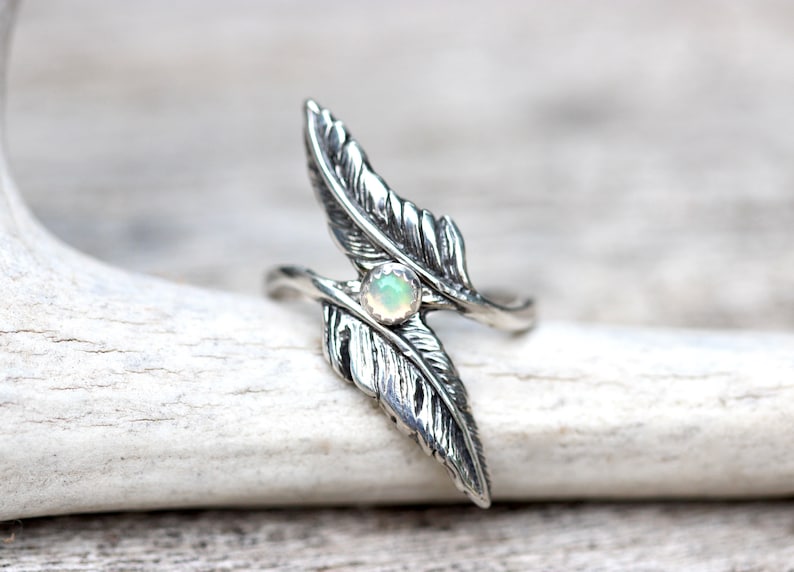 May include: A silver ring with two feather designs and a white opal gemstone in the center.