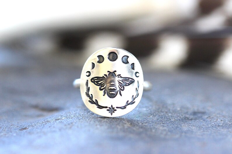 May include: A silver ring with a bee and moon phases design. The bee is in the center of the ring, surrounded by five moon phases.