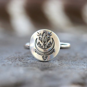 May include: A silver ring with a round, engraved design featuring a crescent moon, a crystal, and an arrow.