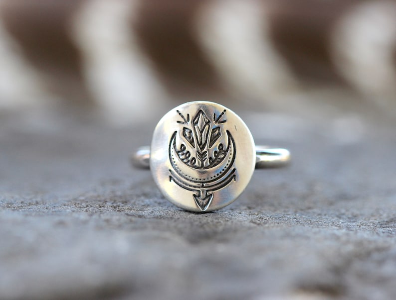 May include: A silver ring with a round, engraved design featuring a crescent moon, a crystal, and an arrow.