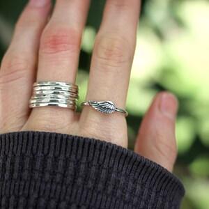 May include: A hand wearing a stack of five silver hammered rings on one finger and a silver wing ring on another finger.