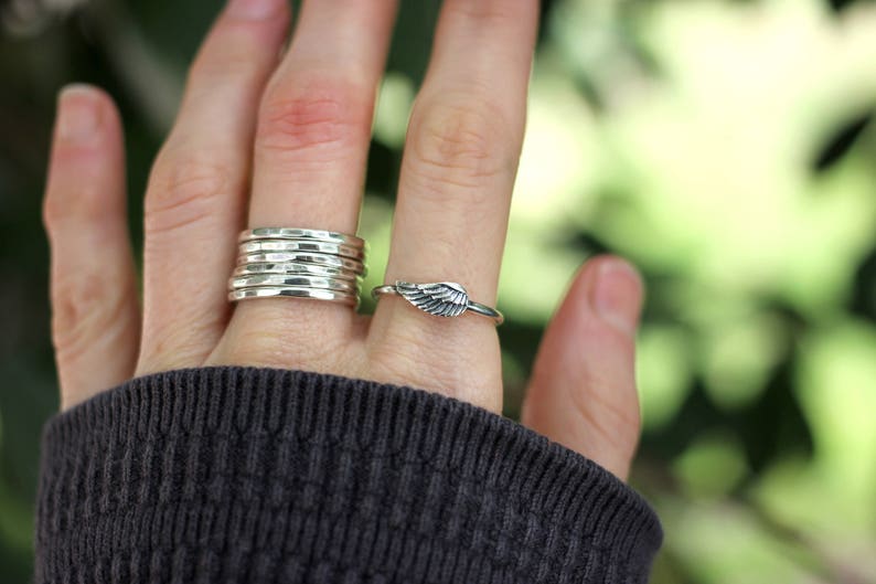 May include: A hand wearing a stack of five silver hammered rings on one finger and a silver wing ring on another finger.