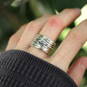 May include: A silver ring with a stack of bands featuring a small wing design. The ring is worn on a person's finger.